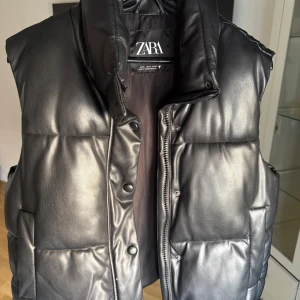 Svart dunväst i skinnlook från Zara - Snygg svart dunväst från Zara i skinnimitation med quiltad design. Västen har hög krage, dragkedja och tryckknappar framtill samt två stora fickor. Perfekt för lager-på-lager och ger en cool vibe till din outfit.