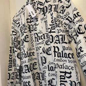 Palace skjorta - Säljer en vit skjorta från Palace Skateboards med svart, stor logotyp- och textprint över hela plagget. Skjortan har klassisk krage, knäppning framtill och långa ärmar. Modellen heter 'Ye Olde But A Goldie Jean' och är riktigt statement.