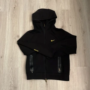 Nike NOCTA svart hoodiejacka - Svart hoodiejacka från Nike NOCTA med gul logga och detaljer. Jackan har hel dragkedja, två fickor med dragkedja och huva. Snygg och sportig look med NOCTA-tryck på ärmen och diskreta detaljer på ryggen. Perfekt för dig som gillar streetwear.
