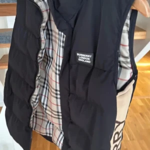 Svart dunväst från Burberry, S - Svart dunväst från Burberry med klassiskt rutigt foder i beige, svart, vitt och rött. Västen har en stor Burberry-logga längs sidan i stickat material och dragkedja framtill. Perfekt för lager-på-lager och riktigt snygg till streetwear eller casual outfits.