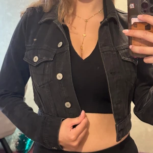 Svart croppad jeansjacka - Säljer en svart croppad jeansjacka med silvriga knappar och klassisk krage. Jackan har långa ärmar och markerad midja, perfekt för att lyfta vilken outfit som helst. Snygg och enkel att matcha med både klänning och byxor.