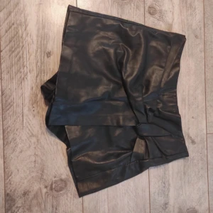 Svart skinnimitation skort från Zara - Snygg svart kortkjol/shorts från Zara i skinnimitation med omlott-look framtill och dragkedja bak. Kjolen har en  cool, edgy vibe och passar perfekt till en statement-outfit. Modellen är figurnära och ger en modern siluett. 