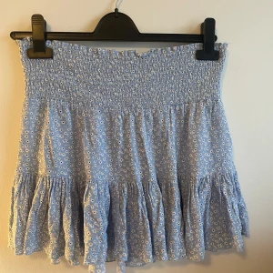 Blommig blå volangkjol från 157 - Supersöt ljusblå kortkjol från 157 med vita små blommor och volanger. Kjolen har smock i midjan för en bekväm passform och är perfekt för vår och sommar. Lätt och luftig känsla, passar till sneakers eller sandaler. Storlek S. Aldrig använd