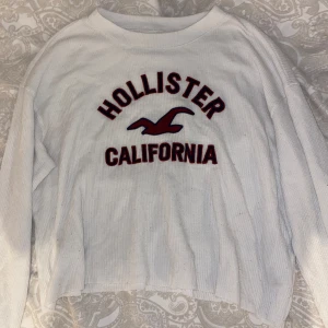 Vit ribbad Hollister tröja - Vit långärmad tröja från Hollister med röd broderad logga och texten 'California' framtill. Tröjan är ribbad och har en avslappnad passform med något croppad längd. Perfekt för en chill och casual look.