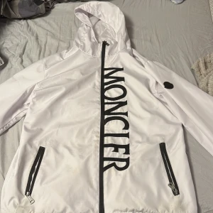 Vit vindjacka från Moncler med huva - Snygg vit vindjacka från Moncler med stor svart logga längs dragkedjan. Jackan har huva, svarta detaljer vid fickor och dragkedjor samt en cool serietidningsetikett på insidan. Perfekt för dig som vill sticka ut med stilren design och streetkänsla. Har en liten fläck på jackan men inget som man lägger mycket märke till.!