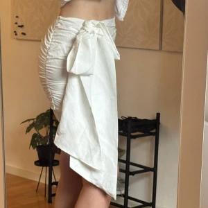 Vit omlottkjol med rosett från Gina Tricot - Supersnygg vit kortkjol från Gina Tricot med omlottkänsla och stor rosett i sidan. Kjolen har rynkade detaljer längs sidorna och är tillverkad i ett lätt, slätt material som känns fräscht till sommaren. Perfekt för dig som gillar trendiga och feminina plagg.