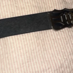 Svart brett bälte med metallspänne - Snyggt svart bälte med brett band och ett coolt, dekorativt metallspänne framtill. Perfekt för att ge outfiten en edgy touch. Bältet är stilrent och passar till många olika looks.