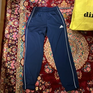 Mörkblå Adidas mjukisbyxor med vita ränder - mörkblå mjukisbyxor från Adidas med vita ränder längs sidorna och klassisk logga på låret. Byxorna har resår och snörning i midjan samt smal passform vid benslut. Perfekta för träning eller chill dagar. Använd ca 5 ggr. Storlek 152. För barn.