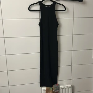 Svart långklänning  - En stilren svart långklänning med hög halsringning och ärmlös design. Klänningen har en rak passform och är perfekt för dig som gillar minimalistisk stil. Enkel att styla med accessoarer för en personlig touch.