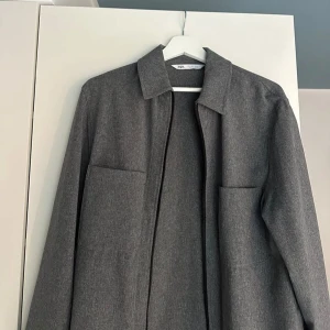 Grå overshirt från Zara, storlek L - Snygg grå overshirt från Zara i storlek L. Jackan har en rak passform, klassisk krage och stora fickor framtill. Perfekt lager-på-lager-plagg med stilren look.