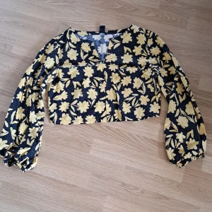 Blommig croppad blus med v-ringning - Supersnygg croppad blus med svart botten och stora gula blommor. Långa puffiga ärmar med resår vid handlederna och en  v-ringning framtill. Perfekt för dig som vill sticka ut med ett coolt blommönster och trendig passform.💕