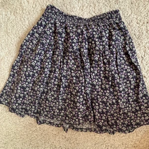 Blommig kortkjol från Zara med inbyggda shorts - Supersöt svart kortkjol från Zara med små blommor i lila, vitt och grönt. Kjolen har resår i midjan och är lätt utställd för en flowy känsla. Perfekt för dig som gillar en romantisk och chill stil. Plus har den inbyggda shorts som är tunna