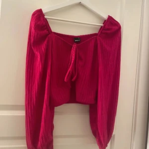 Cerise ribbad blus Amalie topp - Superfin cerise blus från Gina tricot med ribbad struktur och långa puffiga ärmar. Toppen har en snygg knytning framtill och elastisk kant vid axlarna för en offshoulder-look. Perfekt för dig som vill sticka ut med färg och trendig stil.