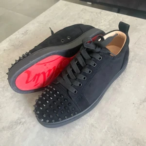 Christian Louboutin svarta sneakers med nitar - Svarta sneakers från Christian Louboutin i mocka med coola svarta nitar på tån och klassisk röd sula. Skorna har snörning och rund tå, samt lyxig läderinsida. Perfekta för dig som vill sticka ut med en edgy vibe.