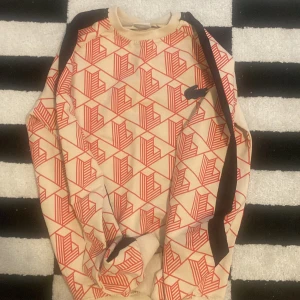 Beige och röd Lacoste sweatshirt - Säljer en beige sweatshirt från Lacoste med rött geometriskt mönster och svarta detaljer längs ärmarna. Tröjan har lång ärm, rund hals och klassisk krokodillogga på bröstet. Perfekt för dig som gillar streetstyle och vill sticka ut.