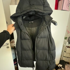 Svart dunväst med huva från SHEIN - Säljer en svart, quiltad dunväst från SHEIN i storlek L. Västen har en stor, mysig huva och dragkedja framtill. Perfekt att slänga över en hoodie för en chill look. Fickor på sidorna och enkel, stilren design.
