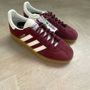 Adidas Handball Spezial vinröda sneakers - Adidas Handball Spezial sneakers i vinrött mocka och textil med vita detaljer och klassiska tre ränder. Skorna har vit plös, vit hälkappa och gummisula. Guldtexten 'Spezial' på sidan ger en extra touch. Följer med beiga snören. Tveka inte att höra av dig!!!
