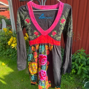 Desigual blommig klänning, M - Färgstark klänning från märket Desigual med svart bas, broderade blommor i rött, gult och grönt samt rosa detaljer. V-ringad, långärmad och med knytband i midjan. 