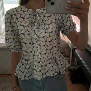 Vit blommig blus med knytband - Supersöt vit blus med blått blommönster, volang nertill och knytband. Endast använd ett fåtal gånger!