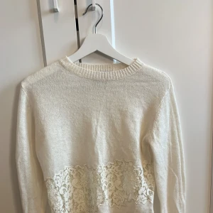 Vit stickad tröja med spets från Zara - Vit stickad tröja från Zara i storlek S. Tröjan har långa ärmar och en bred, dekorativ spetsdetalj i blommigt mönster över magen och ryggen. Mjuk och skön med ribbade muddar vid hals, ärmslut och nederkant. 