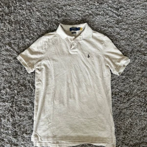 Beige pikétröja från Polo Ralph Lauren - Snygg beige pikétröja från Polo Ralph Lauren med klassisk krage och korta ärmar. Tröjan har broderad logga på bröstet och är gjord i mjuk bomull. Perfekt för en clean och stilren look. Har endast använt den 1 gång. Nypris 1600kr Mitt pris ENDAST 549kr!!!