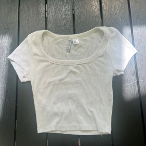 Vit croppad topp från H&M Divided - Superenkel och fräsch vit croppad topp från H&M Divided. Modellen har korta ärmar, bred rundad urringning och är ribbad i materialet. Perfekt till jeans eller kjol för en clean look.