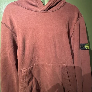 Vinröd hoodie från Stone Island Junior - Säljer en vinröd hoodie från Stone Island Junior med klassisk patch på ärmen. Tröjan har huva, känguruficka och är tillverkad i mjuk bomull. Perfekt för dig som gillar streetwear och vill ha något stilrent men ändå unikt.
