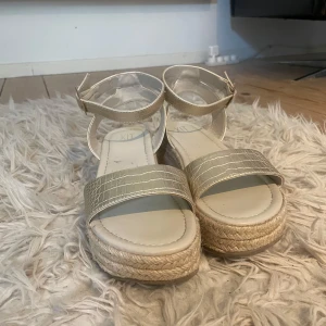 Beiga sandaler med platåsula - Snygga sandaler från XIT i beige med breda remmar i krokopräglat syntetmaterial och justerbart spänne runt ankeln. Platåsulan är klädd i rep för en trendig espadrill-look. Perfekta för varma dagar när du vill ha både stil och komfort.