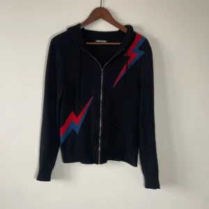 Svart hoodie Zadig & Voltaire med blixt - Cool svart hoodie från Zadig & Voltaire med dragkedja och huva. Tröjan har en röd och blå blixtdetalj på bröstet och sidan, vilket ger en edgy vibe. Tillverkad i mjuk ullblandning, perfekt för dig som gillar statement-plagg med streetkänsla.