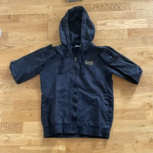 Svart EA7 hoodiejacka med dragkedja - Svart hoodiejacka från EA7 Emporio Armani med korta ärmar, huva och hel dragkedja framtill. Jackan har logga på bröstet och är tillverkad i mjukt bomullsmaterial. Perfekt för en sportig och avslappnad look.