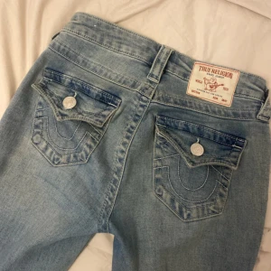 True Religion jeans - Ljusblå jeans från True Religion, modell Becca Mid Rise Bootcut. Klassiska bakfickor med lock och stora silverfärgade knappar, samt tydliga sömmar. Jeansen har en bootcut passform och är tillverkade i jeansmaterial. Perfekta för dig som gillar en avslappnad men snygg stil. Finns några defekter som syns på bilderna, fläcken går säkert bort i tvätten