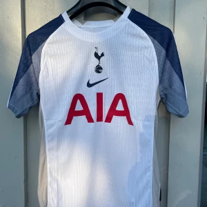 Tottenham Hotspur Nike matchtröja S - Tottenham Hotspur matchtröja från Nike i storlek S. Tröjan är vit med mörkblå ärmar och röda AIA-loggan på bröstet. Tillverkad i lätt och ventilerande Dri-FIT-material, perfekt för fotboll eller supporterliv. Spurs-logga och Nike swoosh framtill.