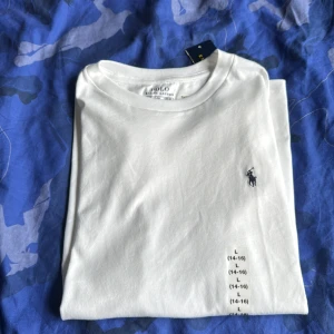 Vit t-shirt från Polo Ralph Lauren - Vit t-shirt från Polo Ralph Lauren med klassisk rund hals och broderad marinblå logga på bröstet. Tillverkad i mjuk bomull som känns skön mot huden. Enkel och stilren design som passar perfekt till jeans eller shorts.