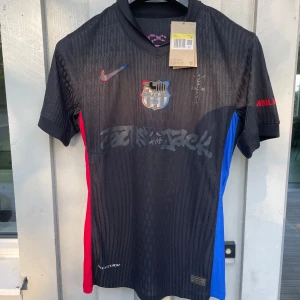 FC Barcelona x Nike svart fotbollströja - Snygg svart FC Barcelona fotbollströja från Nike med coola grafiska detaljer och klubbmärke på bröstet. Tröjan har korta ärmar, röd och blå panel på sidorna och är tillverkad i lätt Dri-FIT material som andas. Perfekt för dig som vill sticka ut i sommar 