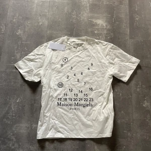 Maison Margiela - Vit t-shirt från Maison Margiela med ikoniskt svart siffertryck och logga på bröstet. Klassisk passform, rund hals och korta ärmar. Tillverkad i mjuk bomull för en clean och stilren look.