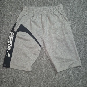 Grå Nike Running shorts med svart detalj - Snygga grå shorts från Nike med svart panel och vit text 'NIKE RUNNING' på vänster ben. De har elastisk midja och två fickor med dragkedja. Perfekta för löpning eller träning, tillverkade i mjuk bomullsblandning för maximal komfort.