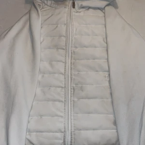 Vit cardigan - Säljer en vit unbrand cardigan med huva. Cardiganen har dragkedja framtill och är lätt vadderad för extra värme. Perfekt att slänga över en hoodie eller tröja när det är kyligt ute. Enkel och stilren look som funkar till det mesta. Den har en liten fläck (3 bilden) men som förmodligen går bort i tvätten