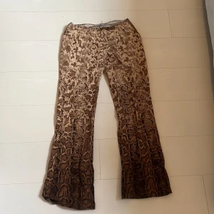 Bruna bootcut byxor med ormmönster - Snygga bootcut-byxor i brunt och beige med ormmönster. Byxorna har dragkedja i sidan och är tillverkade i ett mjukt, lätt material som ger en skön passform. Perfekta för dig som vill sticka ut med ett djurmönstrat plagg.