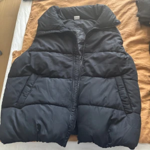 Svart dunväst från Stories M - Säljer en svart, quiltad dunväst från Stories i storlek M (40/42). Västen har hög krage, dragkedja framtill och två snedställda fickor. 