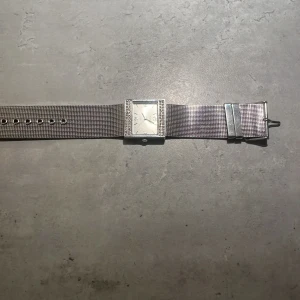 Silvrig fyrkantig klocka Rampage - Stilren klocka från Rampage med fyrkantig urtavla och gnistrande stenar runt ramen. Armbandet är i silvrig mesh och hela klockan har en modern, minimalistisk vibe. Perfekt accessoar för att lyfta din outfit med lite extra bling. Den kommer urladdad 