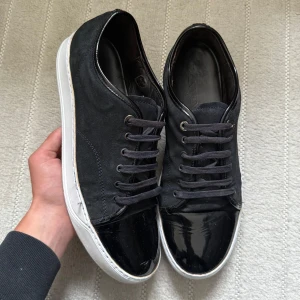 Lanvin Cap Toe Navy - Snygga svarta sneakers från Lanvin med vit sula och svarta skosnören. Skorna har en unik design med blankt lack på tån och mocka på sidorna. Perfekta för dig som gillar stilrena men ändå lyxiga detaljer.
