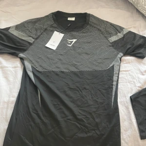 Svart Gymshark t-shirt med mönster - Svart och grå t-shirt från Gymshark med coolt geometriskt mönster och logga på bröstet. T-shirten är kortärmad och har en sportig, tight passform. Perfekt för dig som gillar att träna och vill ha en snygg look på gymmet.