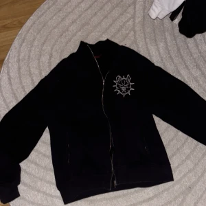 Svart hoodie från Seams  - Svart hoodie från Seams i storlek L. Hoodien har dragkedja, broderad logga på bröstet och stor text på ryggen. Ribbad krage och muddar, samt en liten broderad S-detalj på ärmen. Perfekt för dig som gillar streetwear vibes.