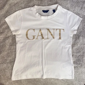 GANT T-shirt  - Vit Gant T-shirt med guldig text. Basic och skön med bra passform. Använd därav lägre pris🫶🏼Storlek 11-12år dvs 152cm. 