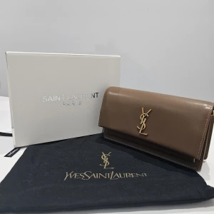 Brun kuvertväska från Saint Laurent - Säljer en lyxig brun kuvertväska från Saint Laurent med ikoniskt YSL-emblem i guld framtill. Väskan är i slätt skinn och har en guldfärgad kedja som axelrem. Insidan har flera fack och magnetlås. Perfekt för dig som vill ha en stilren och exklusiv accessoar.