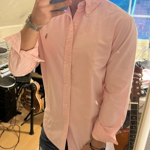 Ljusrosa skjorta från Ralph Lauren - Säljer en klassisk ljusrosa skjorta från Ralph Lauren med button-down krage och broderad logga på bröstet. Skjortan har lång ärm, normal passform och är tillverkad i mjuk bomull. Perfekt för dig som gillar stilrena och tidlösa plagg.