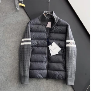 Grå Moncler jacka  med stickade ärmar - Snygg grå pufferjacka från Moncler med stickade ärmar och två vita ränder på varje ärm. Jackan har hög krage, dragkedja framtill och Moncler-logga på vänster ärm. Kroppen är quiltad och glansig medan ärmarna är ribbstickade för en cool kontrast.