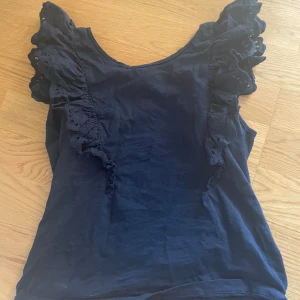 Mörkblå volangtopp Holly & Whyte XS - Supersöt mörkblå topp från Holly & Whyte med volangdetaljer längs axlarna. Ärmlös modell i mjuk bomull, perfekt för dig som gillar romantiska vibes och vill ha något extra till outfiten.