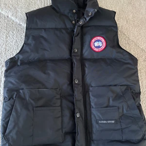 Svart dunväst från Canada Goose - Svart dunväst från Canada Goose med klassisk logga på bröstet. Västen har hög krage, stora framfickor och stängs med både dragkedja och knappar. Perfekt för lager-på-lager och riktigt snygg till streetwear-stil. 