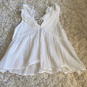 Vit volangblus från Gina Tricot - Supersöt vit blus från Gina Tricot med volangdetaljer vid axlarna och v-ringning med knyt fram. Luftig och flowy modell i tunn bomull, perfekt för varma dagar. Ärmlös och med romantisk känsla som passar till jeans eller kjol. Orginalpris ca 250kr. Pris kan diskuteras. Säljs inte längre 